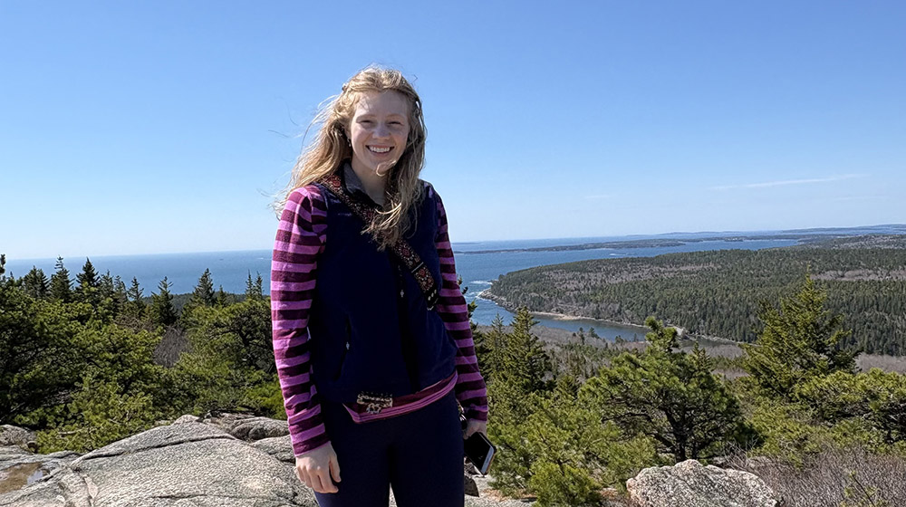 Magdalena Coman standing on top of a rocky hill with blue skies, water, and greenery in the background Magdalena Coman standing on top of a rocky hill with blue skies, water, and greenery in the background